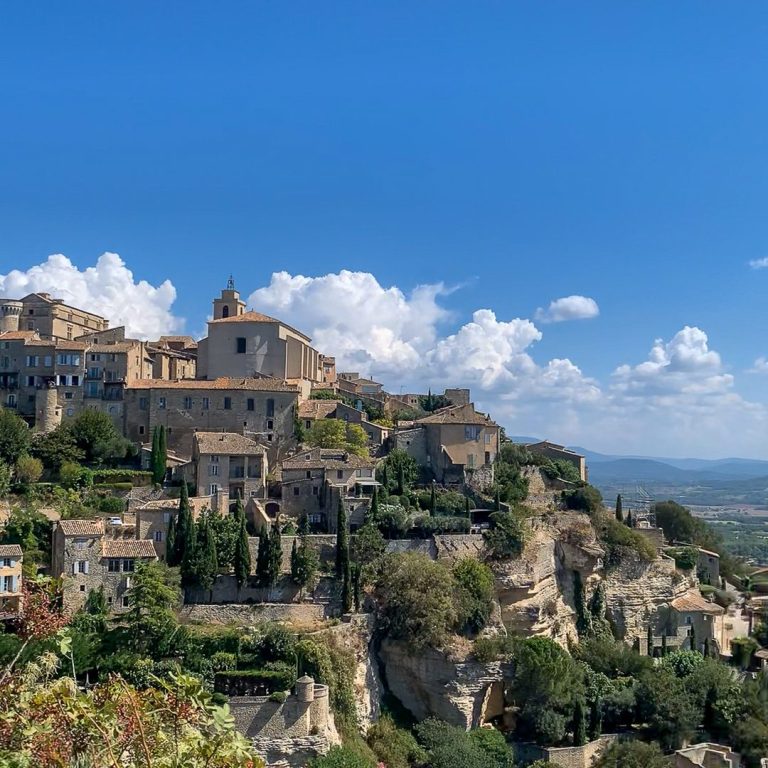 Village médiéval perché sur une colline, entouré de verdure, avec un ciel bleu.