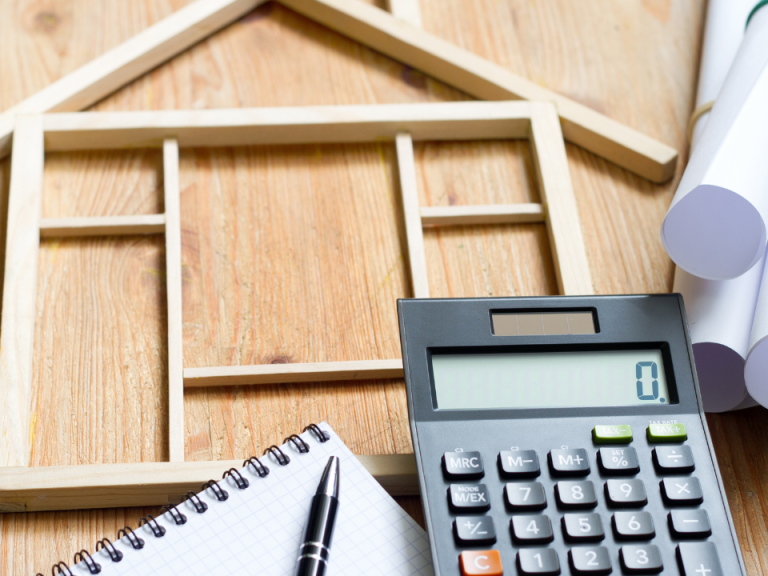 Maquette de maison en bois, calculatrice, carnet et rouleaux de plans sur une table en bois.